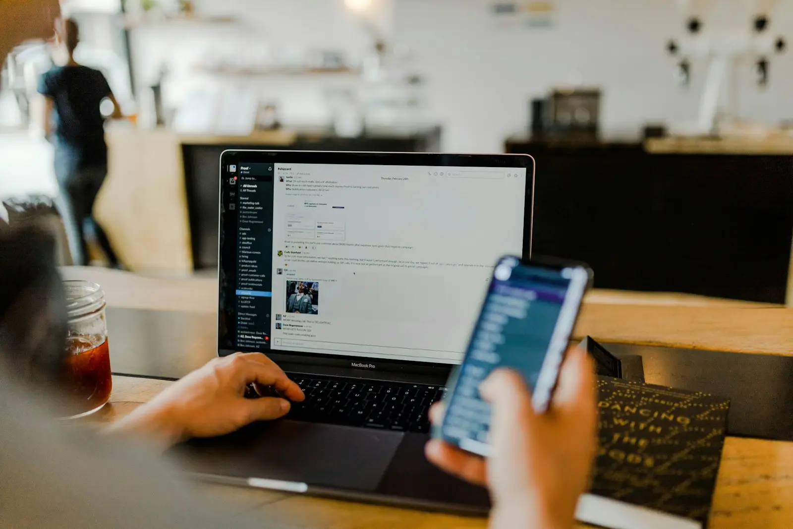 A person at a laptop holding a phone with a chat conversation open on both screens